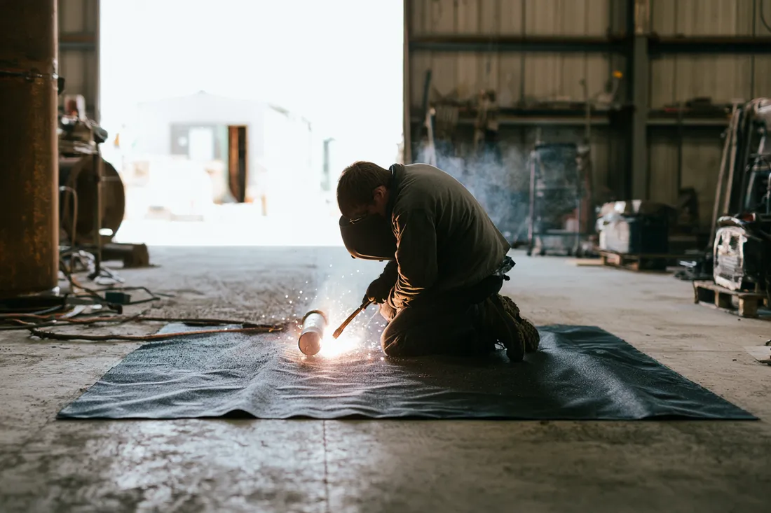 A technician kneeling to weld on a portable fire-resistant mat rolled out over a concrete floor for temporary hot work